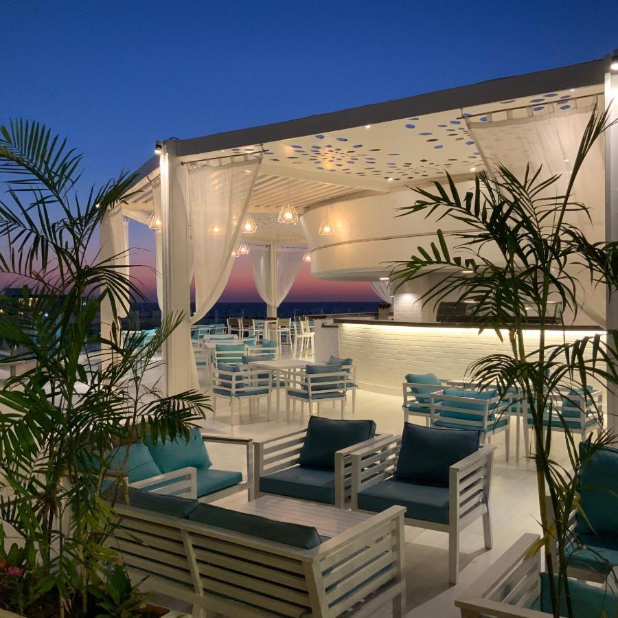 Sunrise Alex Avenue Hotel Aleksandria Zewnętrze zdjęcie The photo shows a stylish outdoor terrace or rooftop lounge area. It features white furniture, including comfortable seating with teal cushions. The space is adorned with draped sheer curtains, providing a relaxed and elegant atmosphere. Lush green p