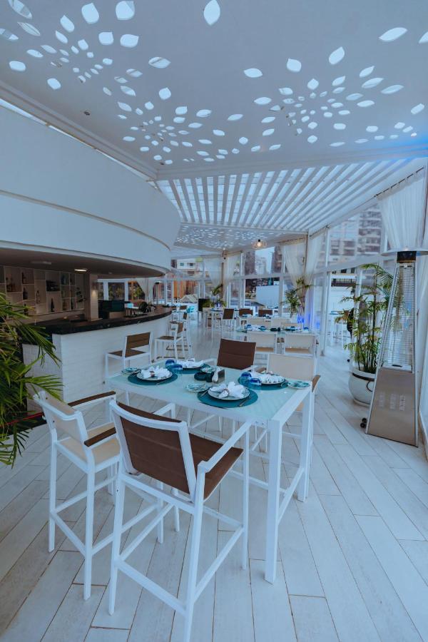 Sunrise Alex Avenue Hotel Aleksandria Zewnętrze zdjęcie The photo shows a bright and modern restaurant interior. The space features white and light-colored furniture, including tables with a combination of white and brown chairs. Each table is elegantly set with plates, utensils, and decorative elements. 