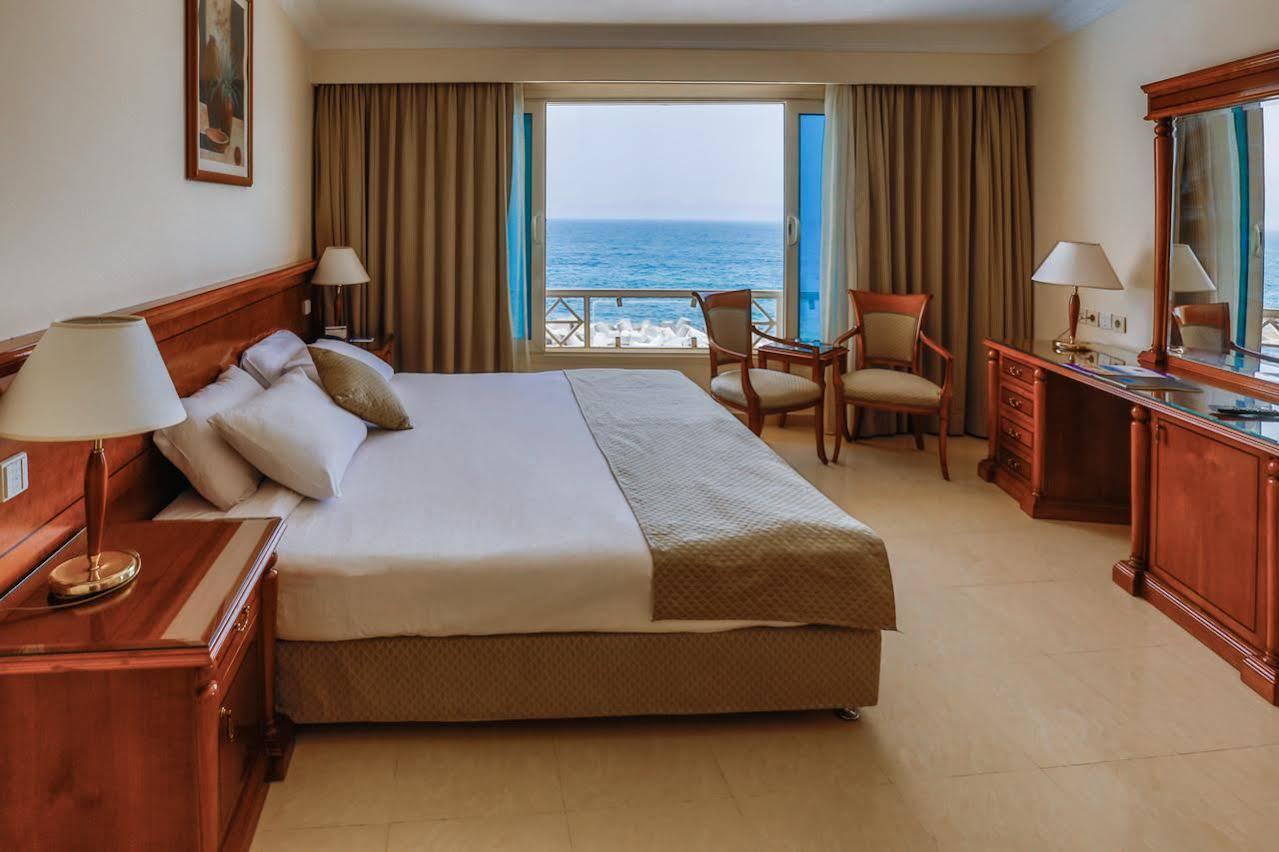 Sunrise Alex Avenue Hotel Aleksandria Zewnętrze zdjęcie The photo shows a hotel room featuring a large bed with white linens and decorative pillows. There are two wooden chairs positioned near a window, which offers a view of the ocean. The room has beige walls and light-colored curtains that frame the wi
