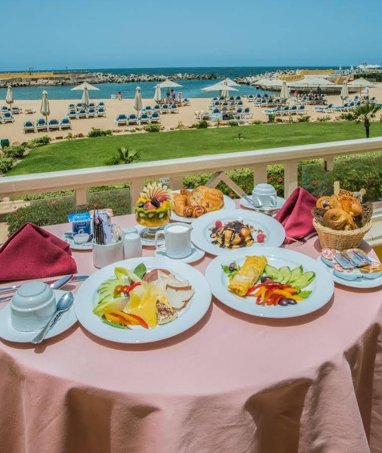 Sunrise Alex Avenue Hotel Aleksandria Zewnętrze zdjęcie The photo shows a beautifully set breakfast table on a balcony with a scenic view of a beach. There are several plates of food arranged attractively. One plate features an assortment of fruits, including slices of melon, berries, and other colorful f