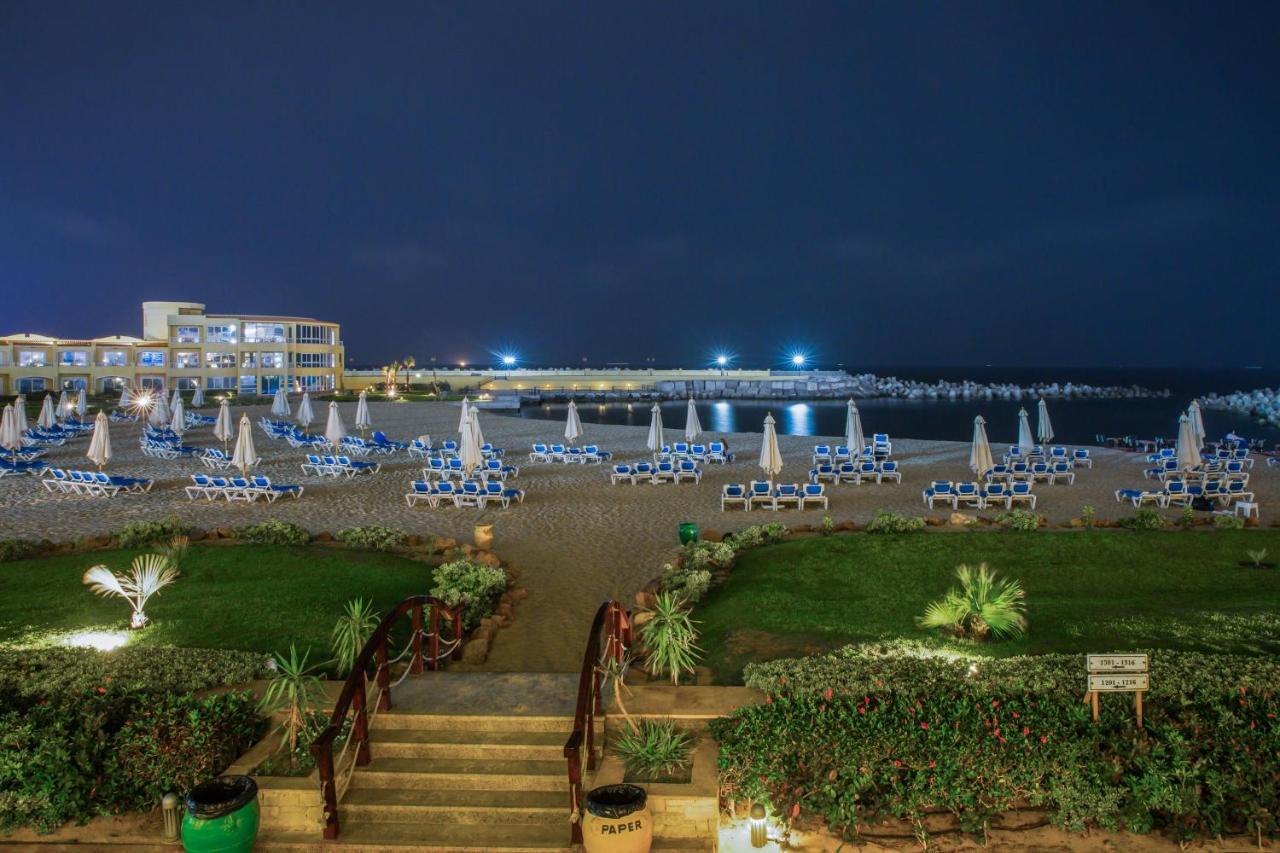 Sunrise Alex Avenue Hotel Aleksandria Zewnętrze zdjęcie The photo shows a beach scene at night. In the foreground, there are steps leading down from a grassy area lined with plants. The beach is filled with numerous lounge chairs, all arranged with umbrellas, suggesting a relaxed atmosphere. In the backgr