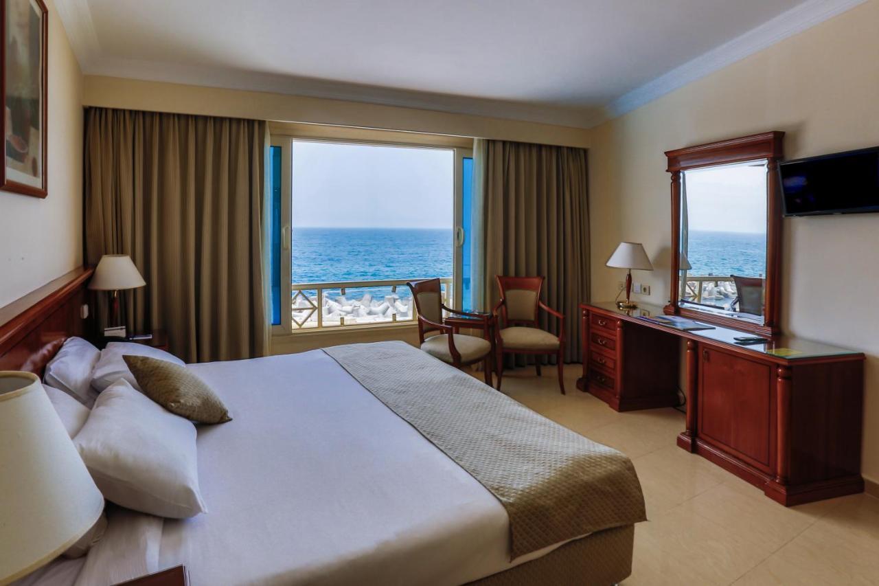 Sunrise Alex Avenue Hotel Aleksandria Zewnętrze zdjęcie The photo shows a hotel room with an ocean view. The room features a large bed with neutral-colored bedding and pillows. There are curtains covering the windows, allowing plenty of light to enter. Beside the bed, there is a wooden desk and a chair po