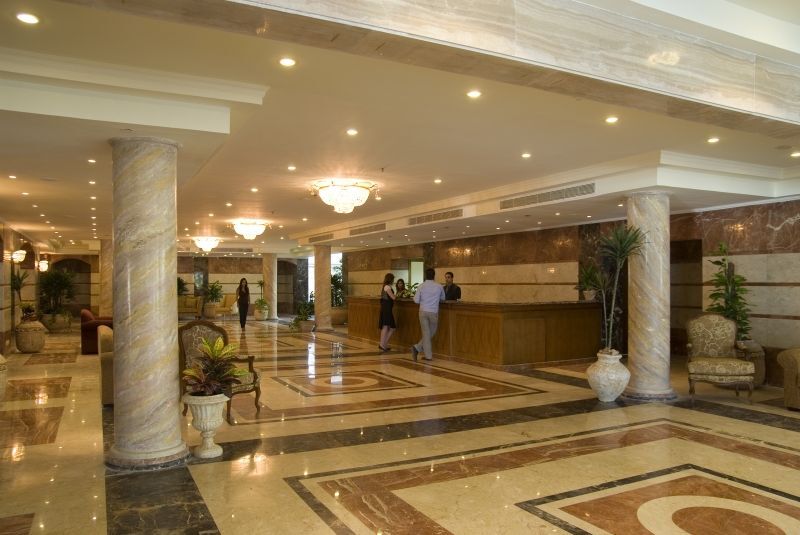Sunrise Alex Avenue Hotel Aleksandria Zewnętrze zdjęcie The photo shows the interior of a spacious lobby, likely in a hotel. There are marble floors with intricate patterns, elegant lighting fixtures, and tall pillars. In the center, there is a reception desk where staff members are attending to guests. S
