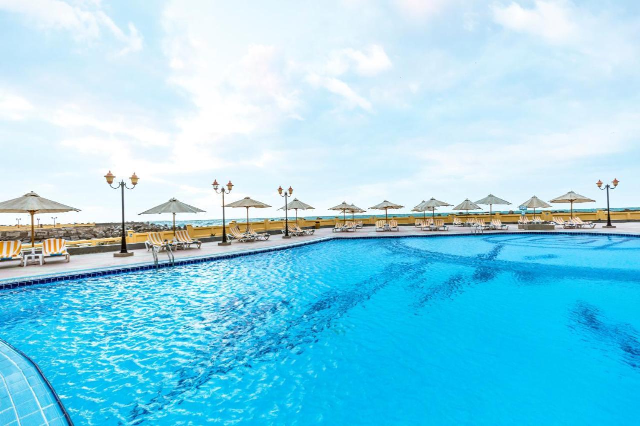 Sunrise Alex Avenue Hotel Aleksandria Zewnętrze zdjęcie The photo shows a large swimming pool with crystal clear blue water. Surrounding the pool are several sun loungers and umbrellas, providing shade for relaxation. In the background, there appear to be more lounge chairs and additional umbrellas, sugge
